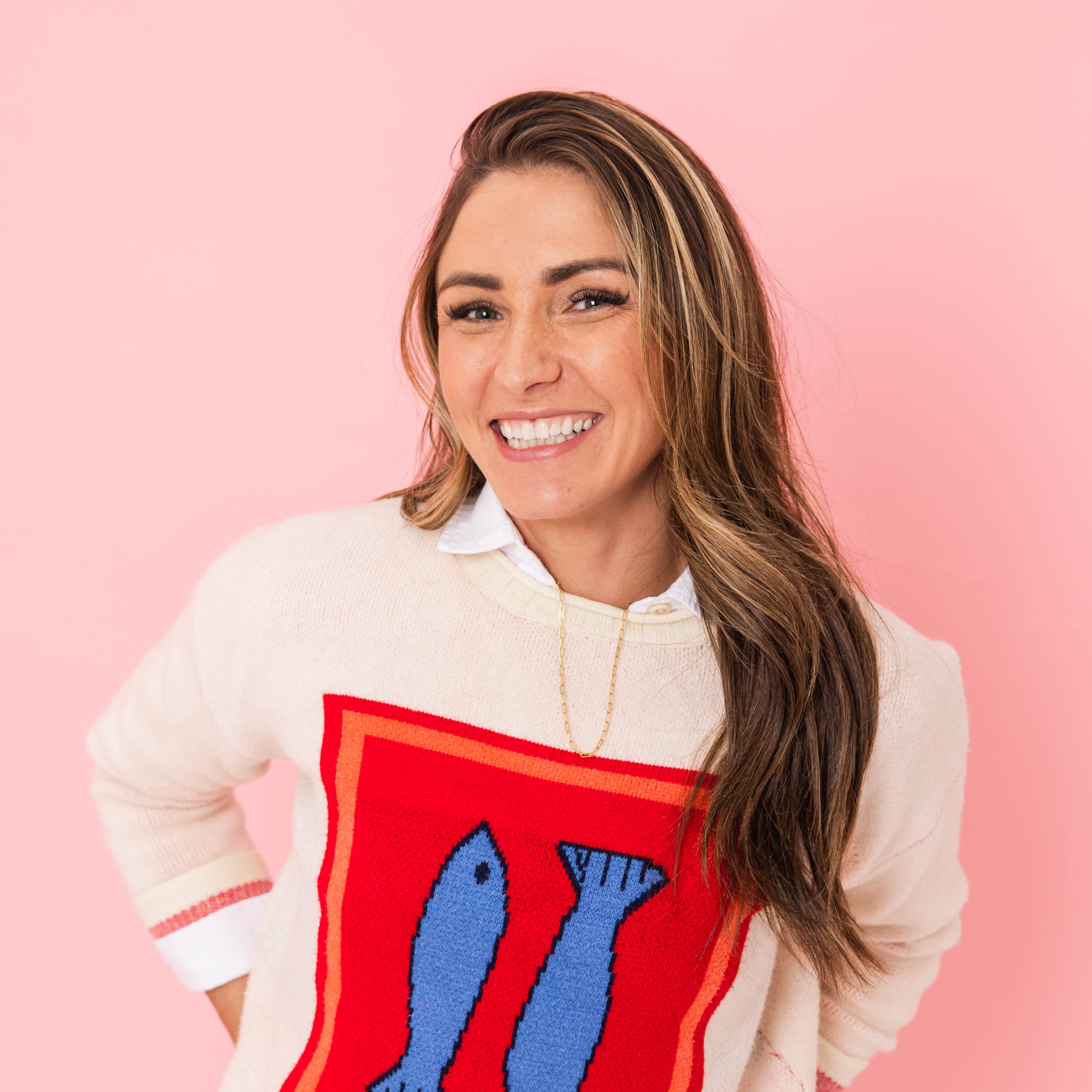 Frannie, founder of Punch Needle Addict stands smiling at the camera with a pink solid background, wearing a white sweater with a red square and 2 blue fish on it.